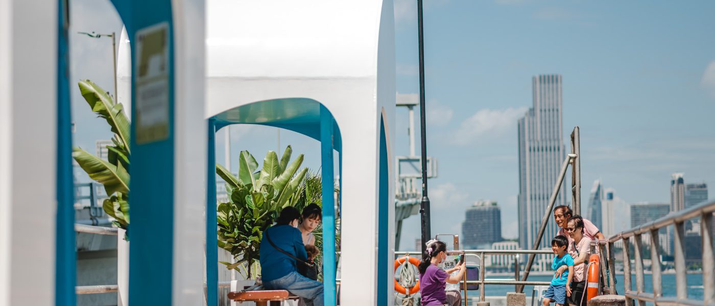 Seating area featuring white and blue sheltered structures along a waterfront with people gathered.
