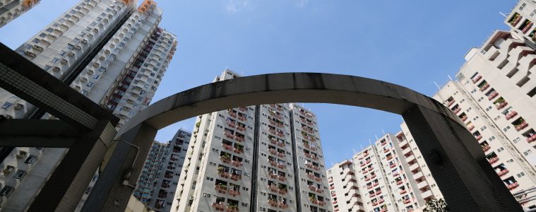 Towering residential blocks surround a curved concrete arch beneath a clear blue sky in a dense urban courtyard.