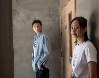 A young man and woman stand beside a wooden doorway in an unfinished concrete interior, facing the camera.