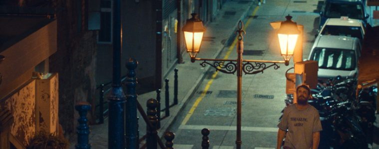A man stands on a steep Macau street beneath ornate lamps, with parked cars, railings and tiled pavements around him.
